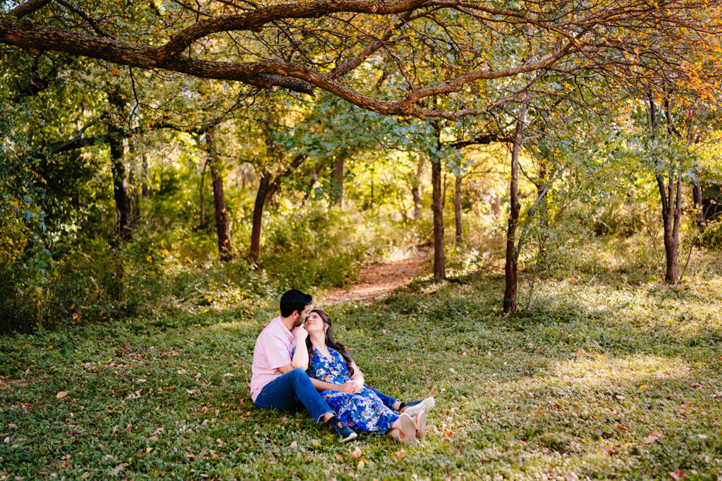 Couple sitting in the grass cuddling near a sunlit trail during their Dallas engagement session at Reverchon Park.