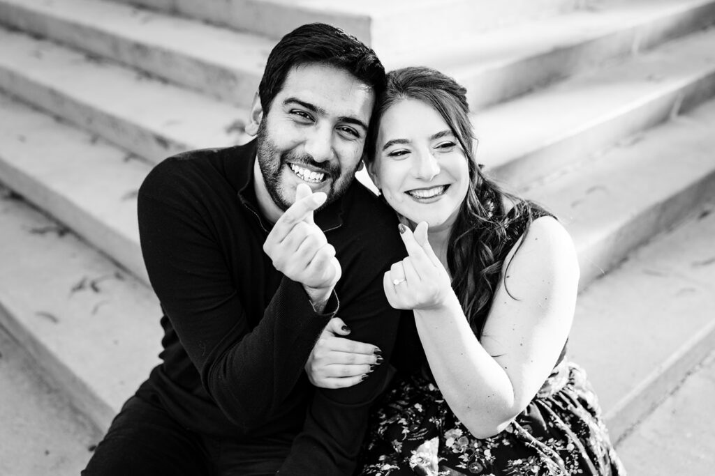 Black and white photo of couple making heart hands while sitting on the steps out front of the meyerson symphony center