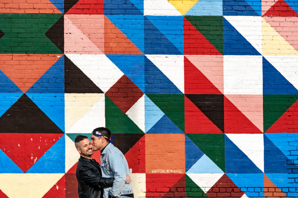 men stand near colorful wall while one kisses the others cheek