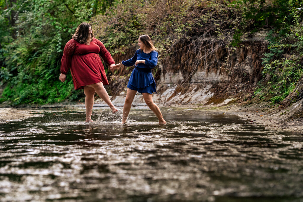woman hold hands while walking in a creek together and playfully kicking water